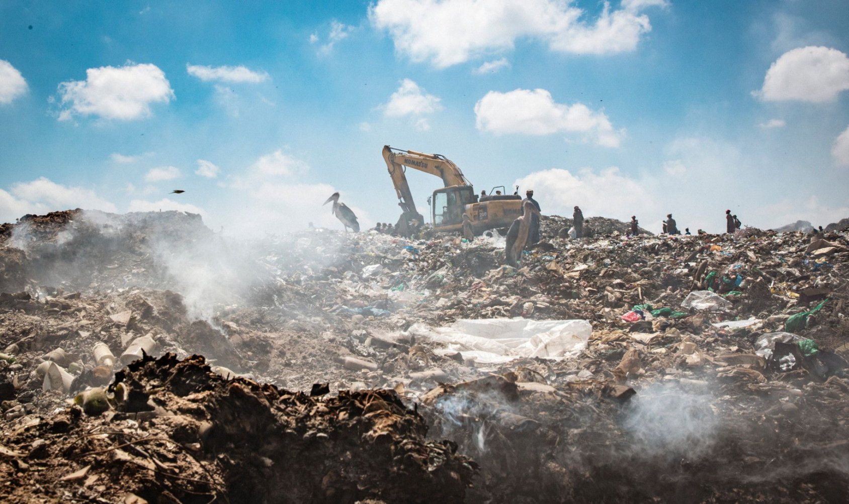 A landfill site with trash all around and small smoking fires. A construction vehicle is on a pile and a bird perches on the trash.