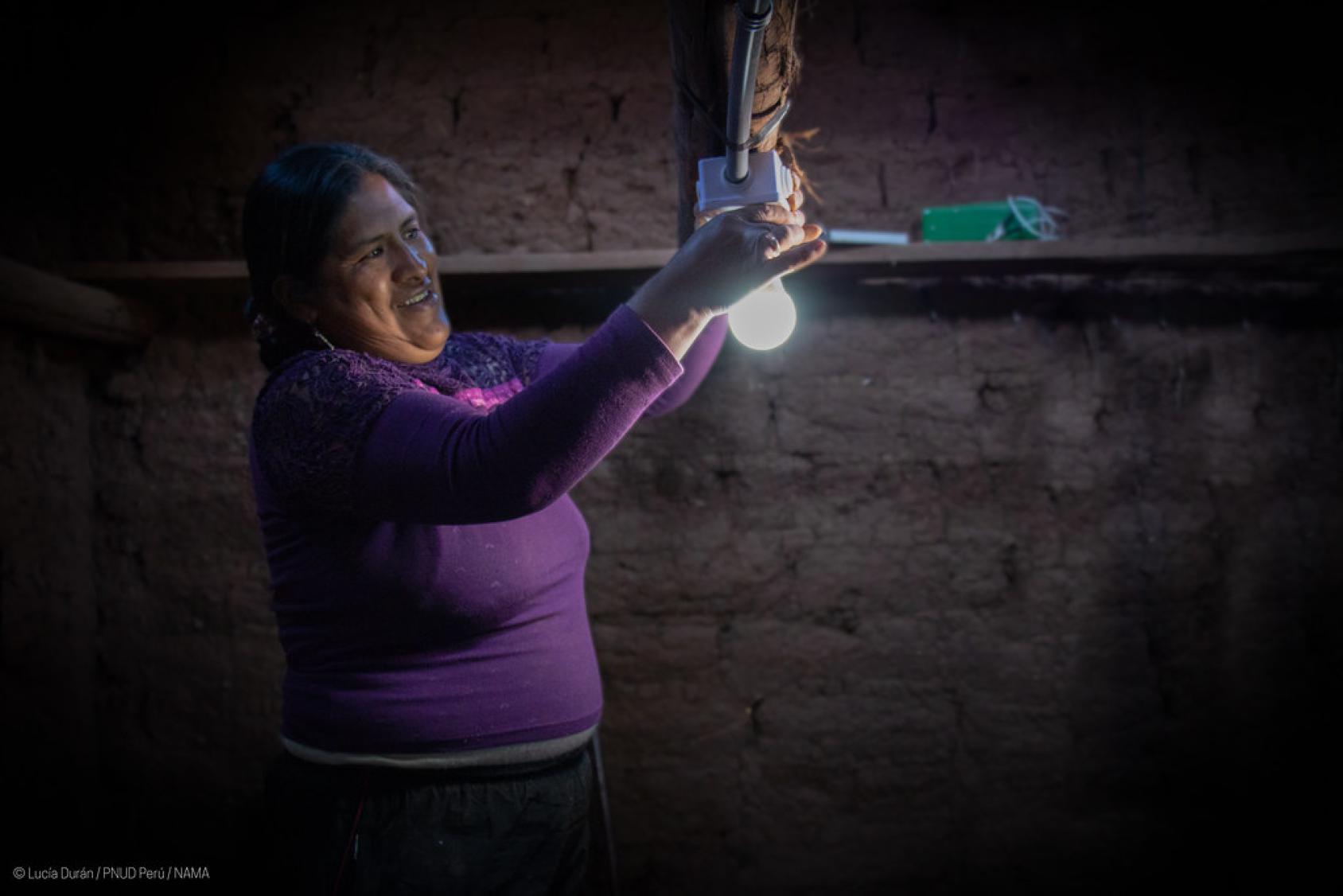 A woman in a purple sweater screws on a light bulb in a dark house