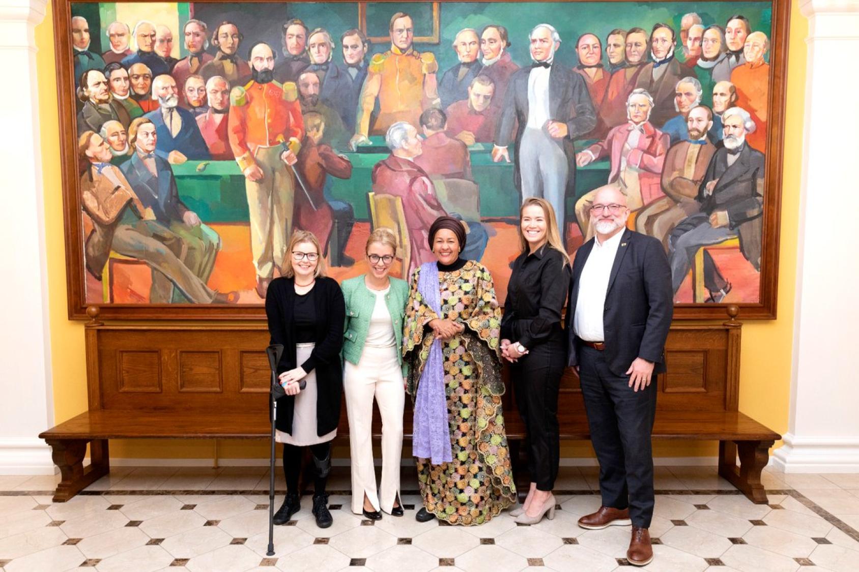 UN deputy chief stands with parliamentarians in Iceland, in front of a colorful bright painting.