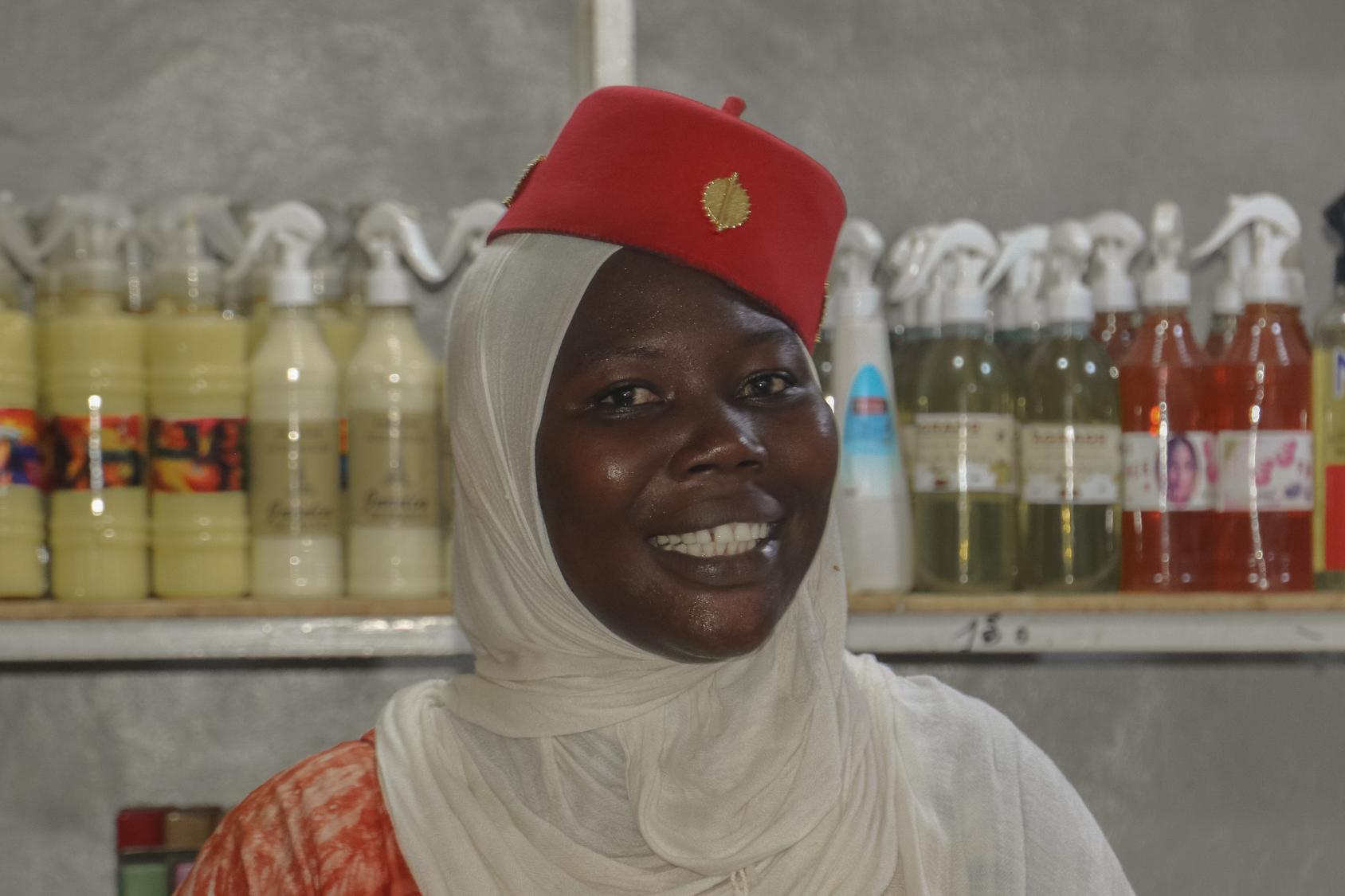 A woman in a white headscarf and red cap stands in front of shelves of beauty products