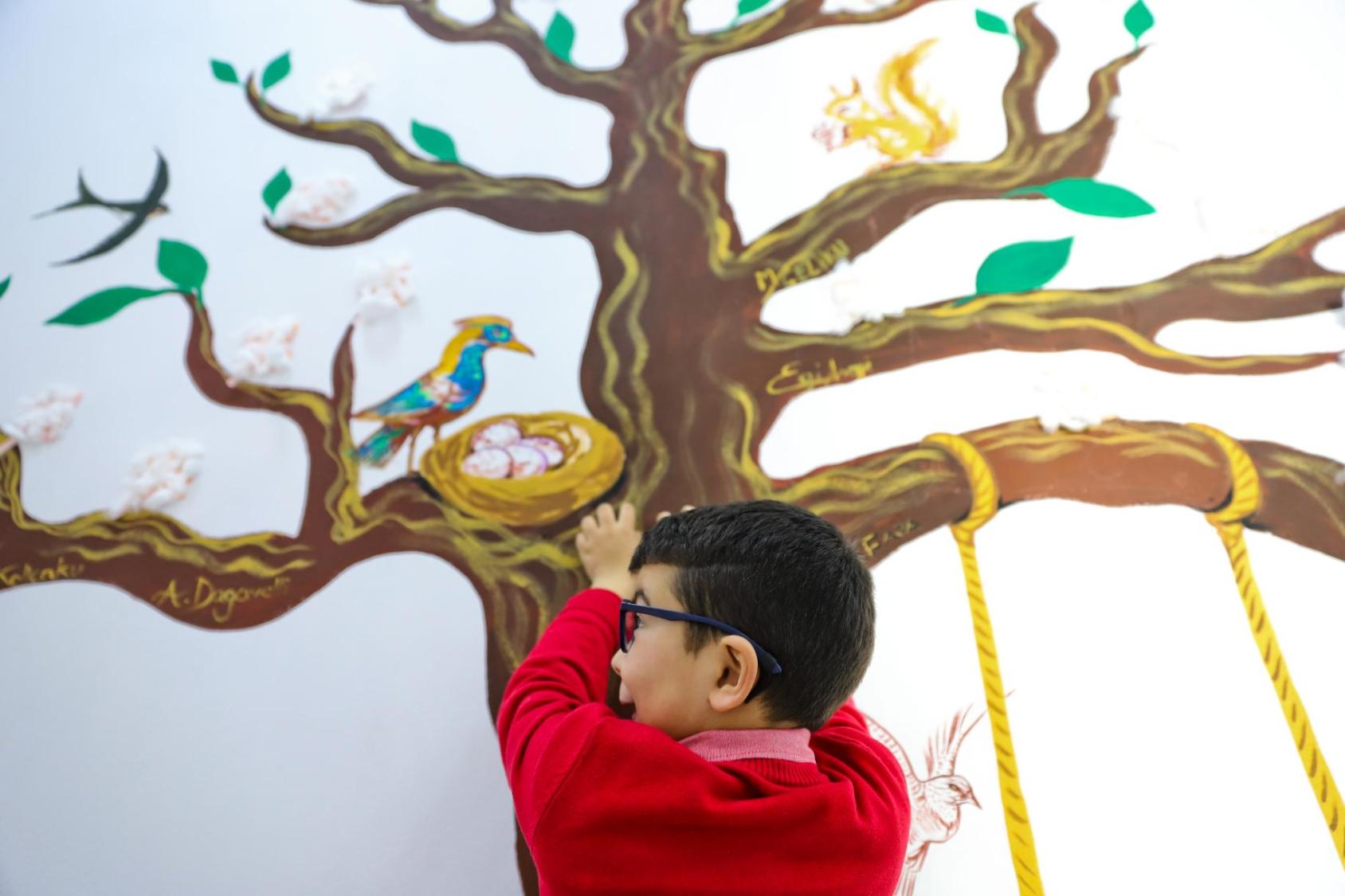 A child in a red sweater stands against a wall with a tree painted on it