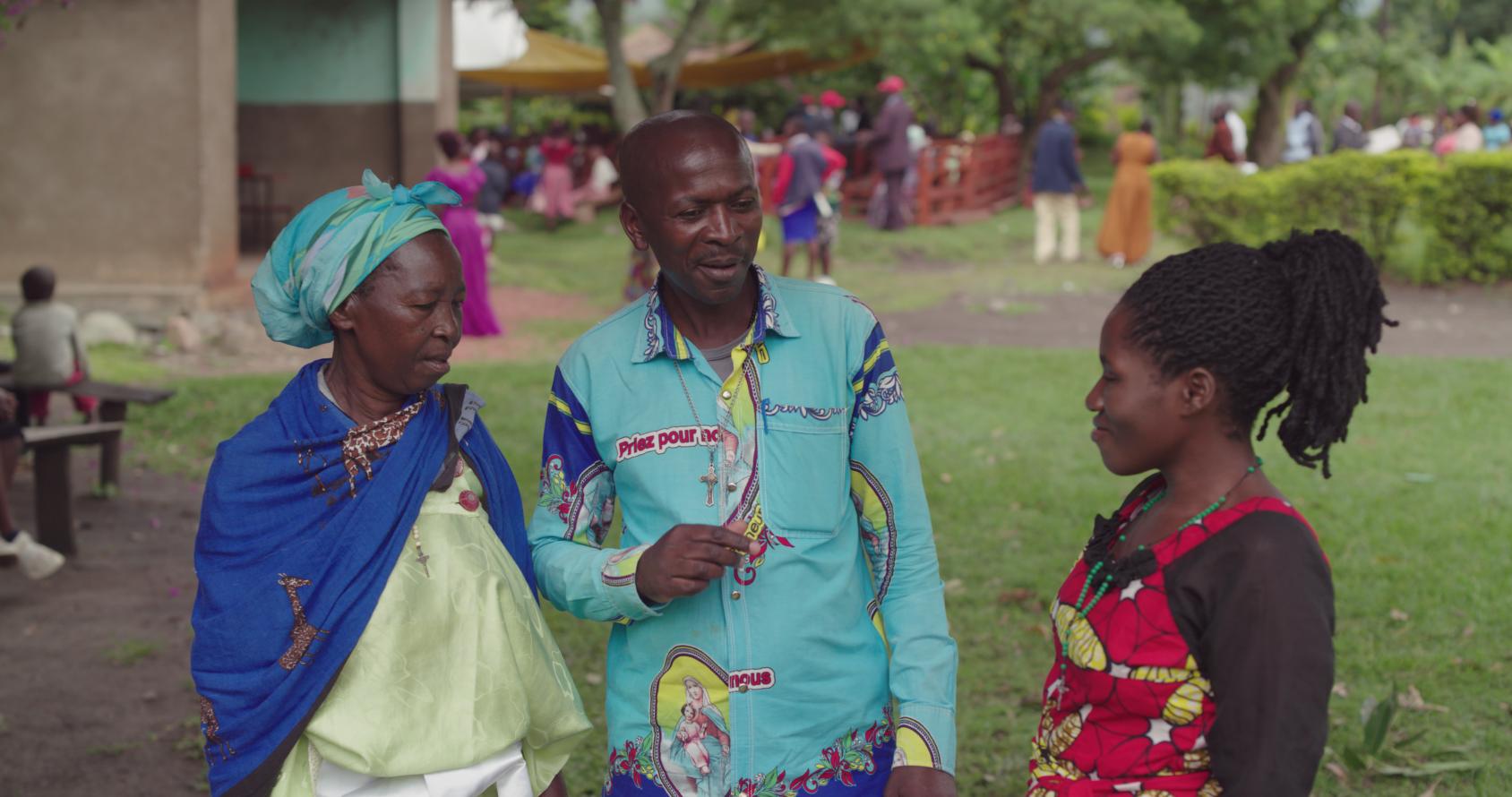 Two women in colourful clothes speak to a man standing in between them in a bright blue shirt in Kasere district in Uganda.