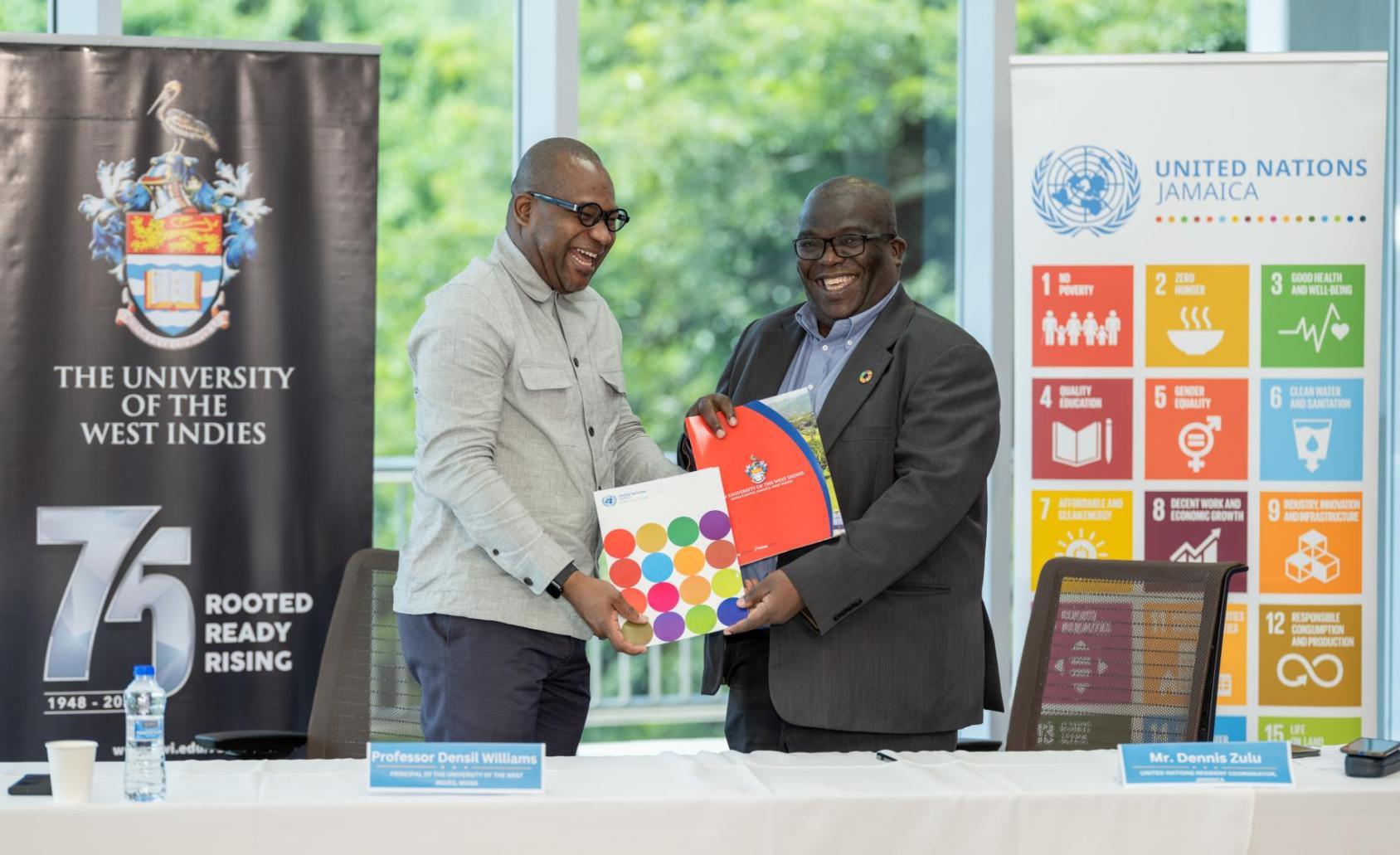 Two men in suits smiling and shaking hands while holding colourful reports on SDGs