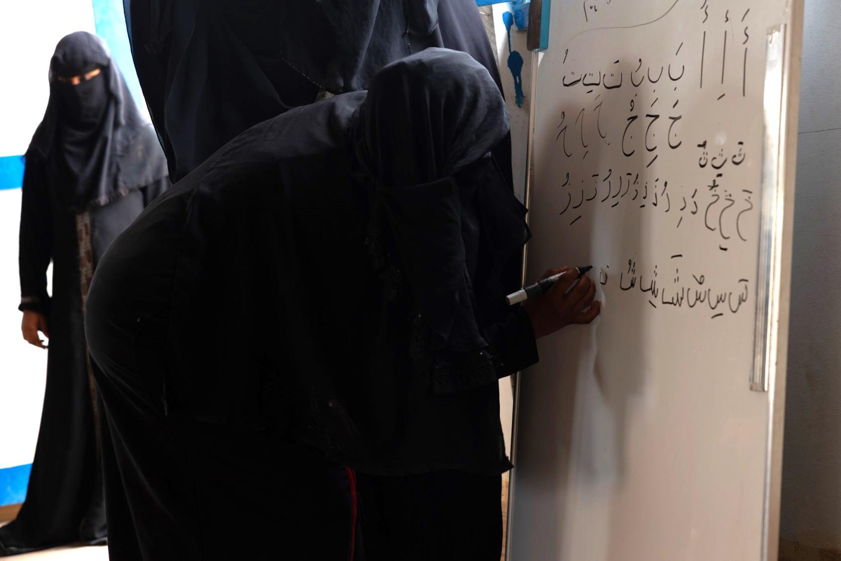 A woman in a black abaya, fully covered from head to toe, writes with a marker on a white board while another woman in grey looks on.