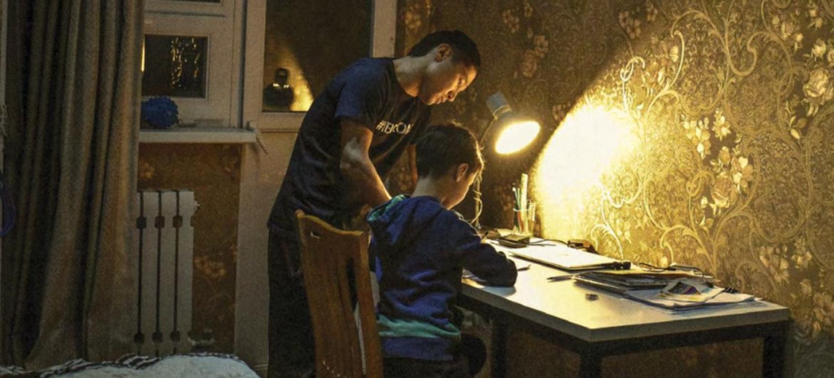 Отец склонился над сыном и помогает ему с домашним заданием