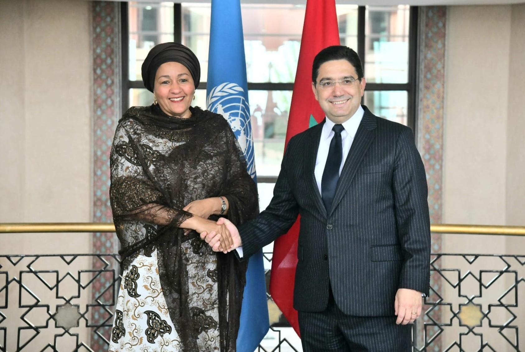 A woman in a black and grey clothing and headscarf shakes hands with a man in a black suit and spectacles against the background of two flags