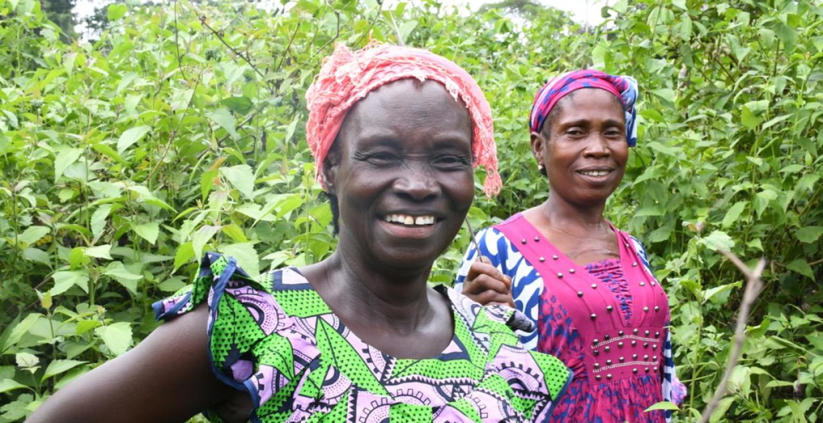 Two Liberian women in brightly coloured clothes stand next to each smiling, in front of green bushes