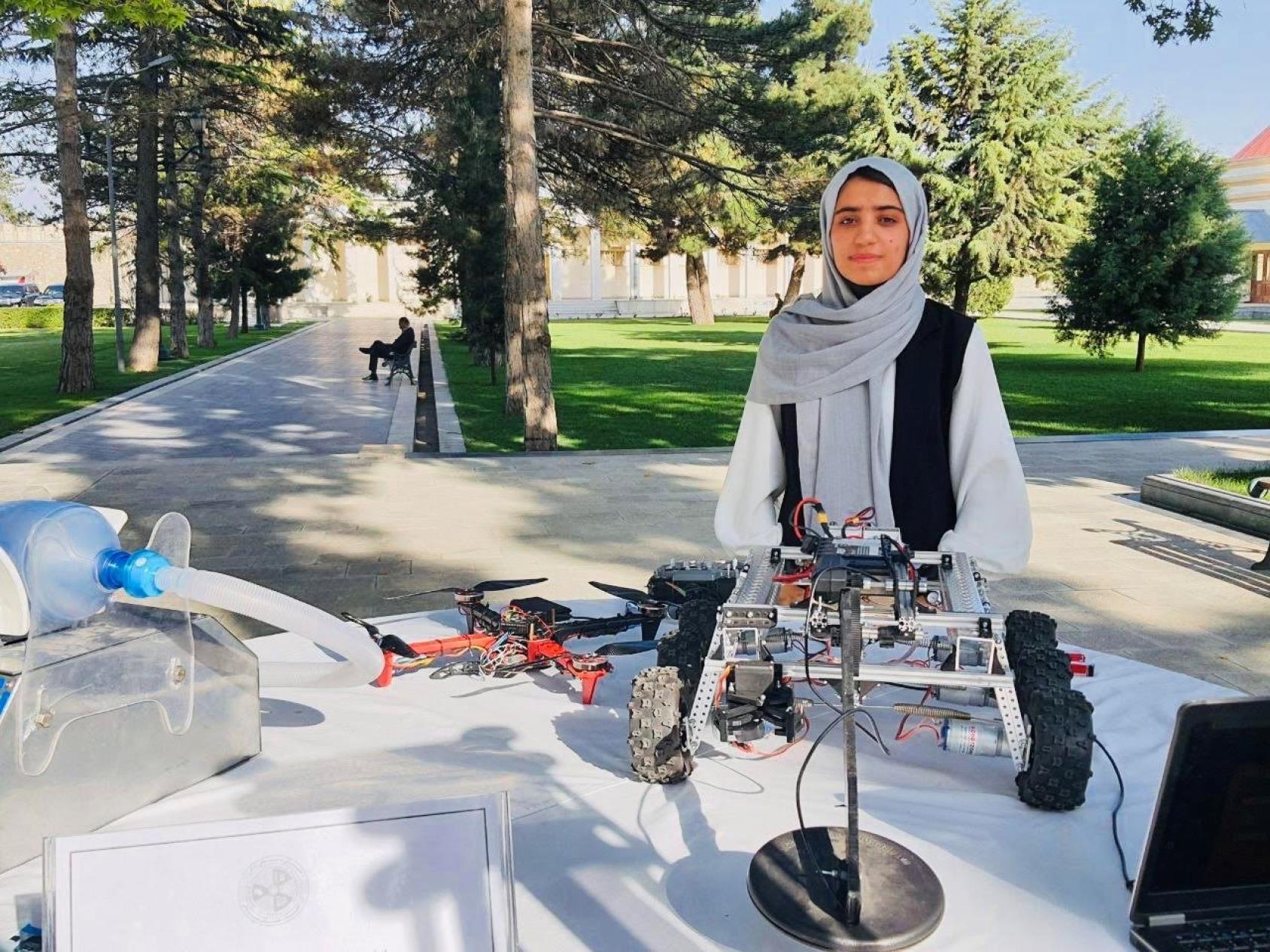 A girl in a black dress and grey headscarf stands in front of a table where a robot model is assembled