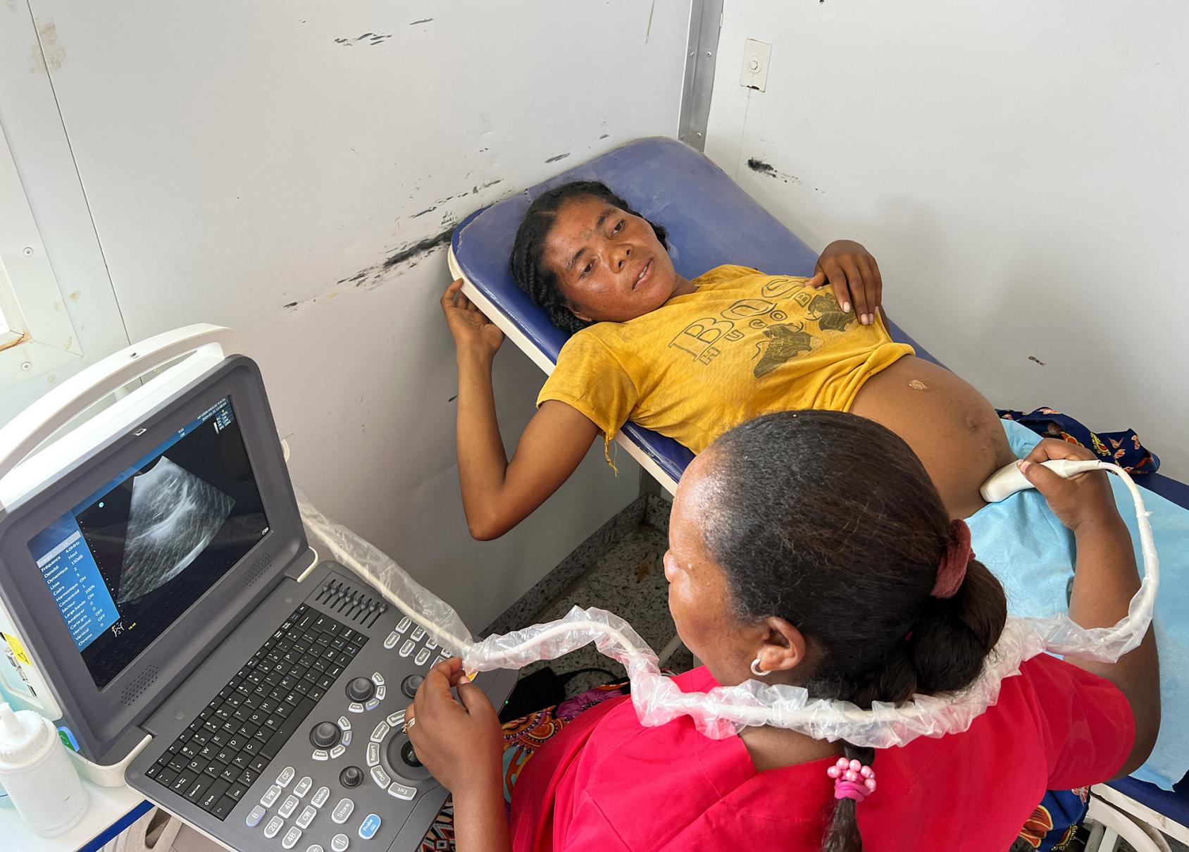 A pregnant woman in a yellow dress lies on a bed while a doctor in a red shirt scans her stomach with a medical device.