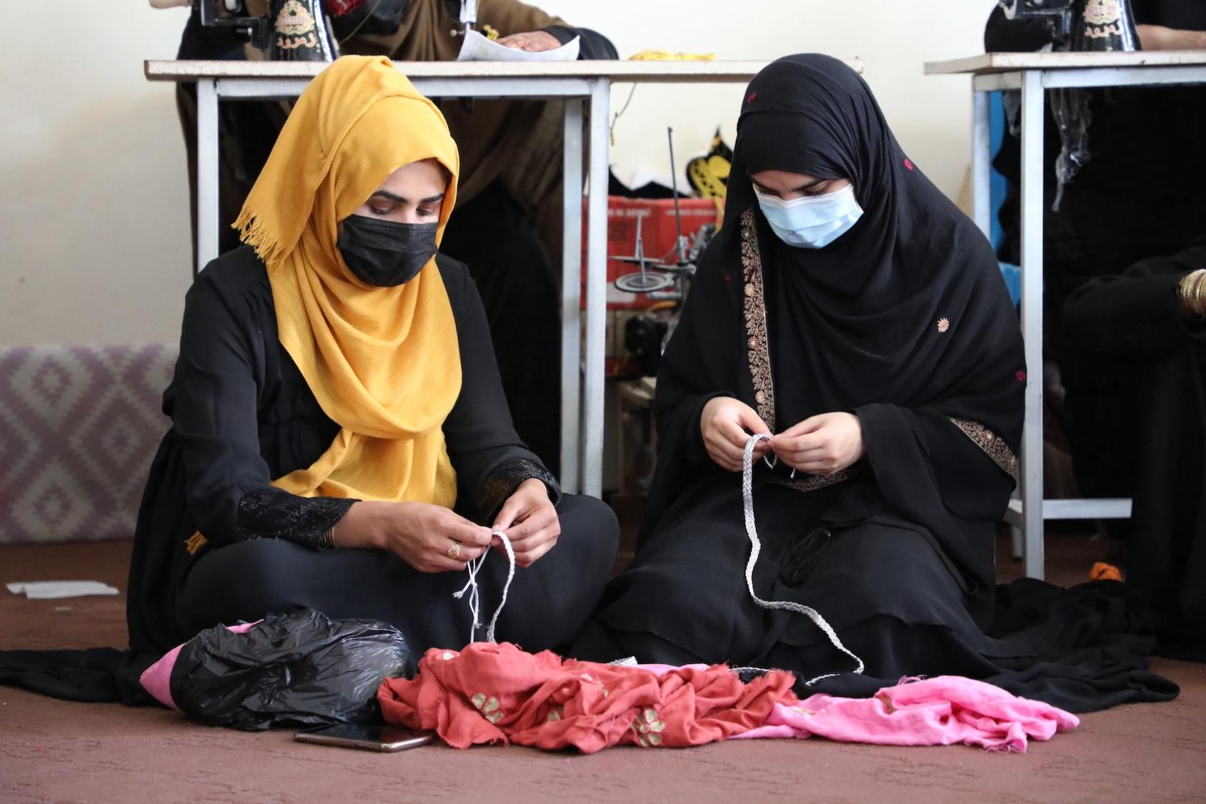 Women engaging in weaving, they are seated on the floor wearing masks and their heads are covered