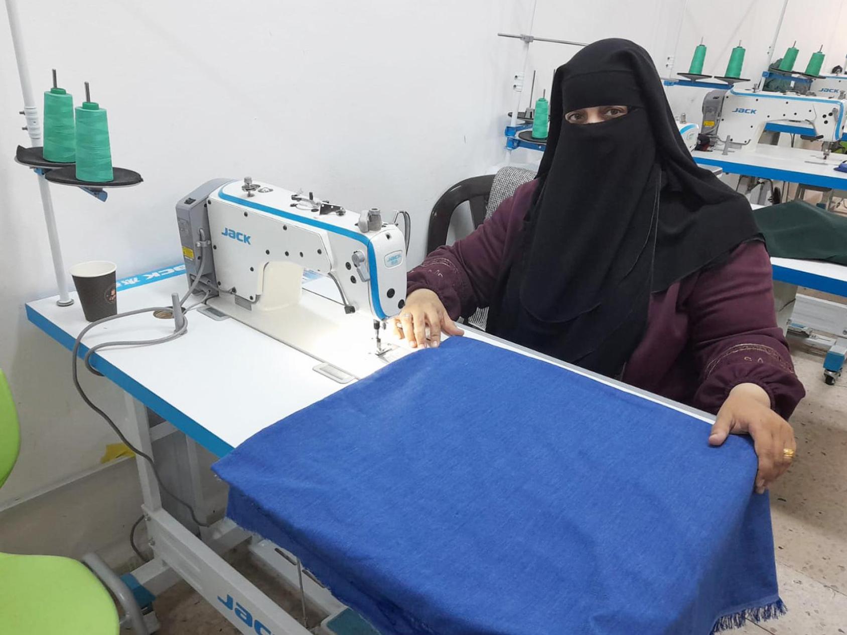 A woman in a dark purple abaya dress with a black headscarf sits in front of a weaving machine with a blue cloth spread in front of her.