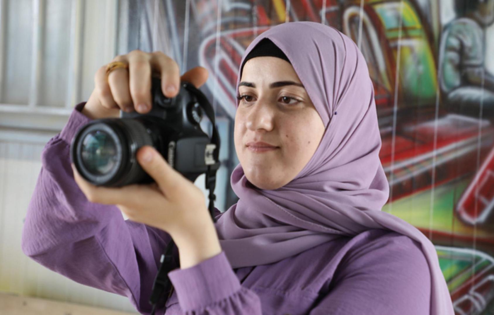 A woman in a lilac dress and headscarf looks through a camera as she takes a picture.