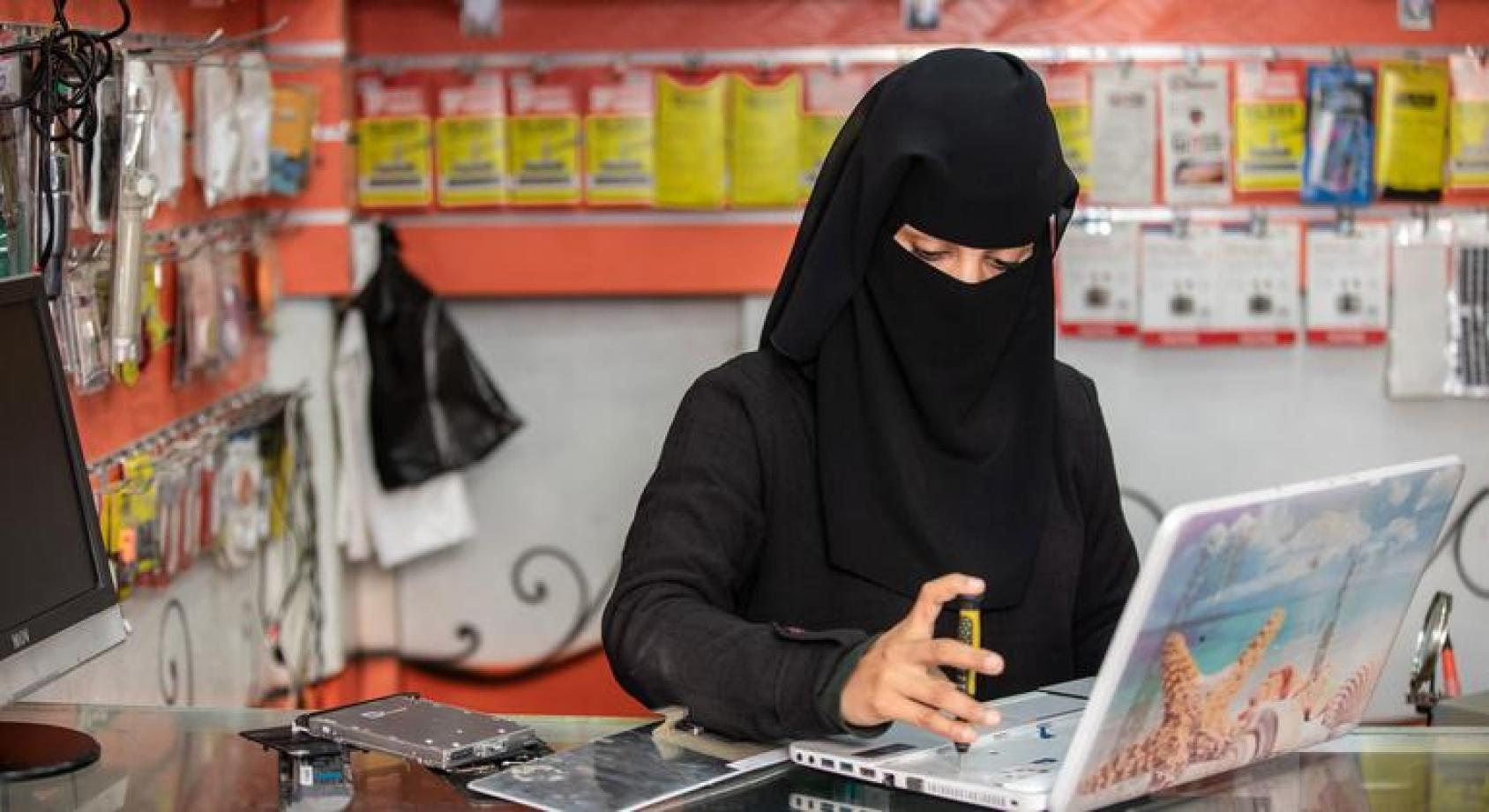 Женщина с покрытым лицом, сидит работает за компьютером в магазинчике техники