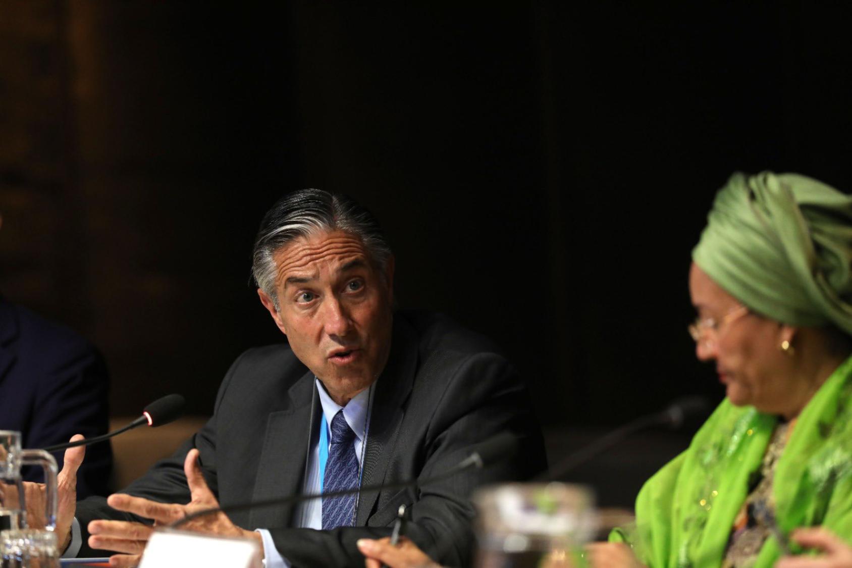 A man in a dark suit and blue tie speaks into a microphone while a woman in a green dress and headscarf listens keenly to him.