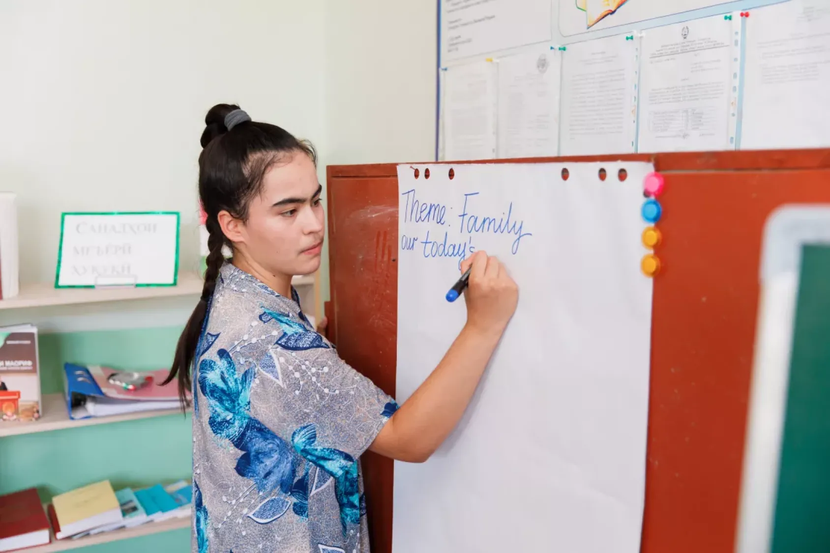 A teacher in a blue shirt and long braid writes on a paper board in a classroom.