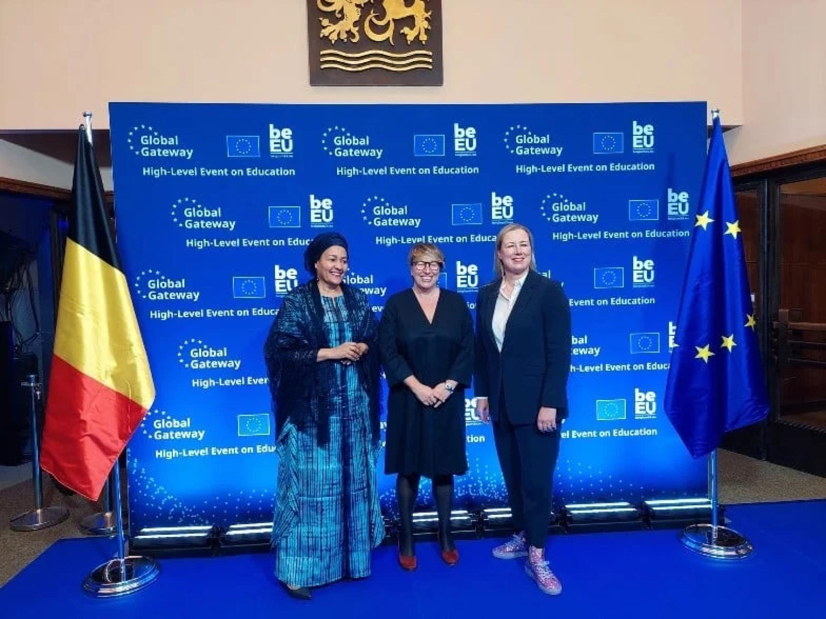 Three women stand in front of a backdrop that has the EU logo. On either side of the backdrop is the EU flag and Belgian Flag