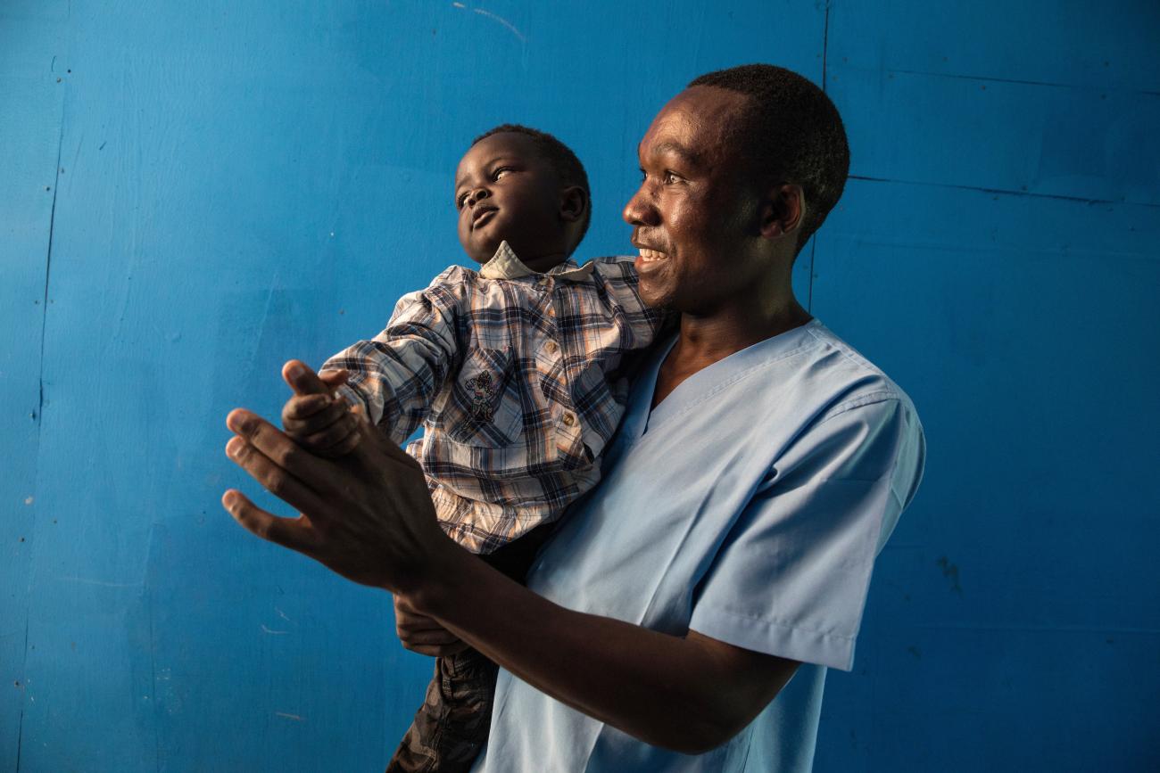 Un homme photographiéde profil sur un fond bleu tient un enfant dans les bras et regrade devant lui en souriant.