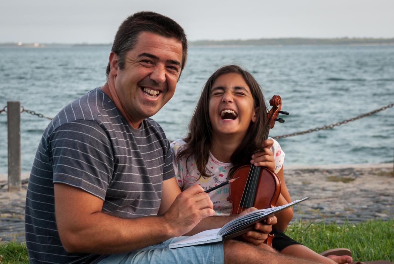 Un hombre con un cuaderno y un lápiz, junto a una niña con un violín, ríen juntos. En el fondo se aprecia el mar.