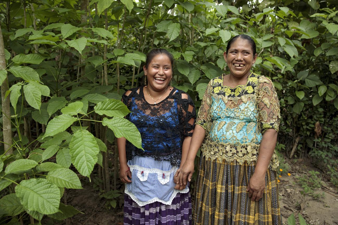 Deux femmes issues d’une communauté autochtone d’Amérique latine se tiennent debout, côte à côte, face caméra, au milieu d’une forêt, et arborent un large sourire. 