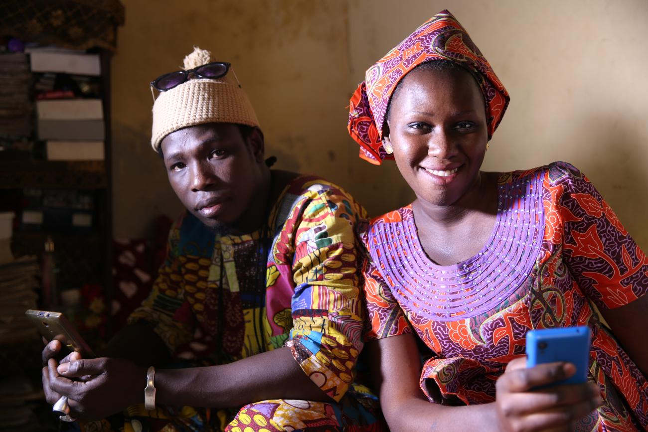 Une femme et un homme sénégalais vêtus d'habits traditionnels sont assis côte à côte et regardent l'objectif de la caméra en souriant et en tenant leur téléphone portable dans les mains.