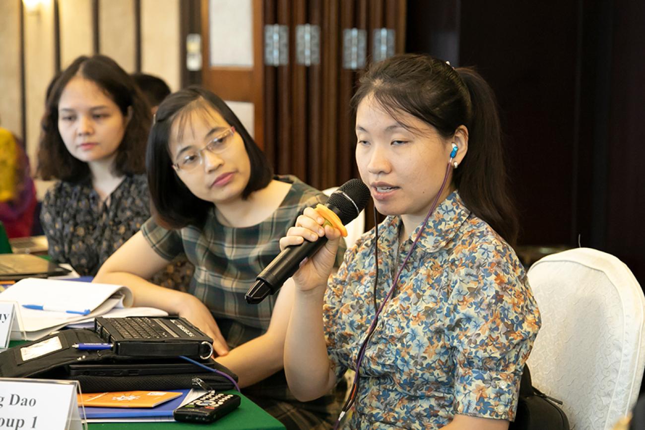 Une jeune femme asiatique assise à une table à côté de deux autres jeunes femmes s'exprime à un microphone.