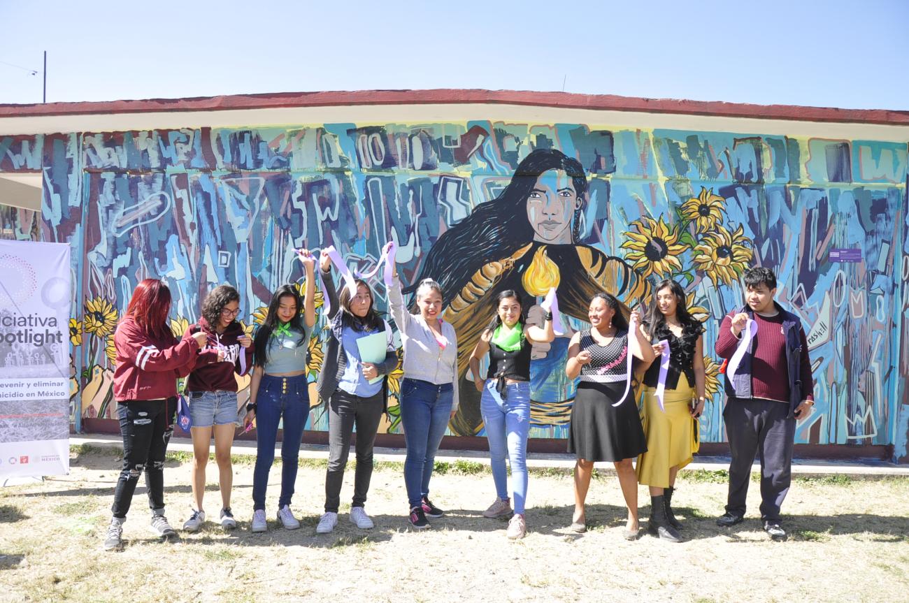 Plusieurs jeunes filles et un jeune homme se tiennent debout fièrement, face à la caméra, devant une peinture murale représentant une femme aux longs cheveux bruns.