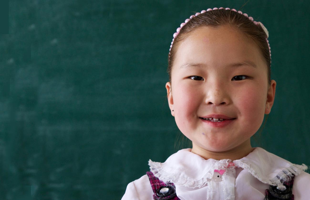 Show a little Mongolian girl smiling at the camera as she stands in front of a blank chalkboard.