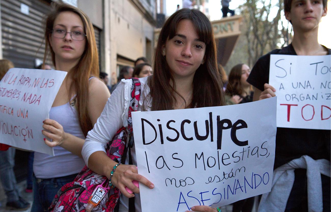 Tres manifestantes jóvenes sostienen carteles de protesta. El cartel de la joven en el centro versa: "Disculpe las molestias, nos están asesinando".