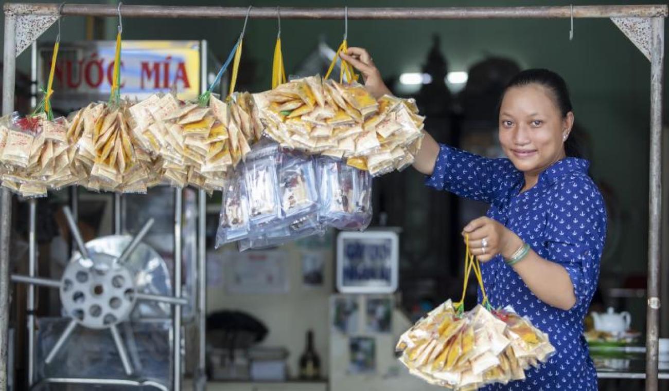 Una mujer con un vestido azul está comprando en un puesto del mercado. Lleva una bolsa de condimentos en la mano y mira a la cámara con una sonrisa.
