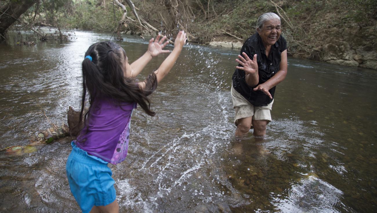 Allizon Stefany Escobar, de cuatro años, salpica a su bisabuela riendo, Conzuelo Flores, mientras ambas están paradas en aguas poco profundas, en un río cerca de su casa, en el distrito de Cayo.