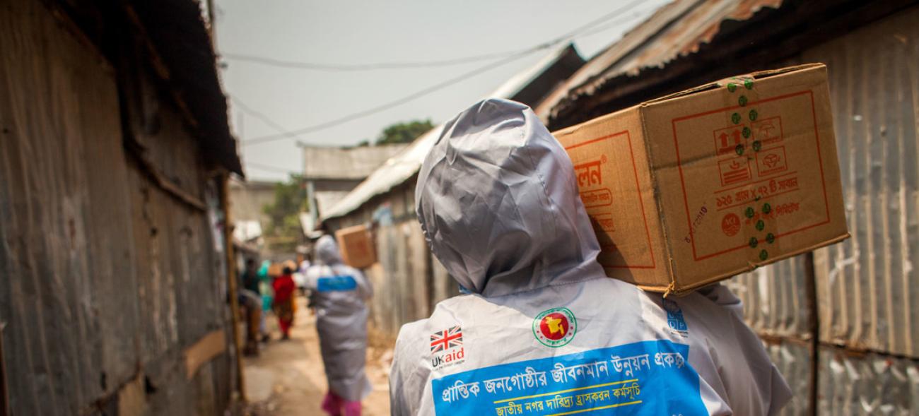 Une personne, dos à la caméra, marche le long d’un sentier séparant deux rangées de cabanes. Elle porte un carton sur l’épaule droite et est vêtue d’un T-shirt sur lequel figurent des inscriptions en bengali et le logo d’UK Aid.
