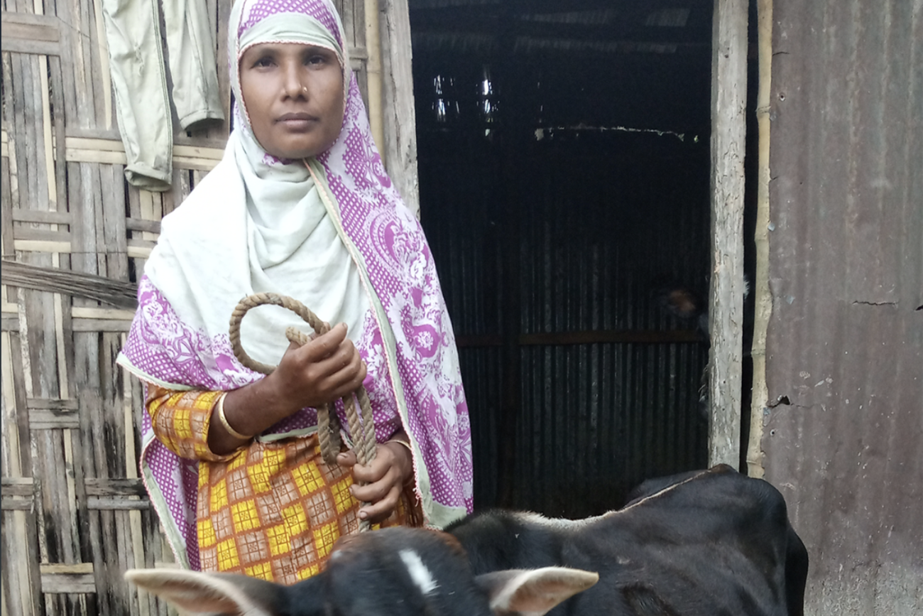 Una mujer, que lleva un velo y sostiene una vaca con una cuerda, se encuentra frente a una puerta de madera, mirando a la cámara.