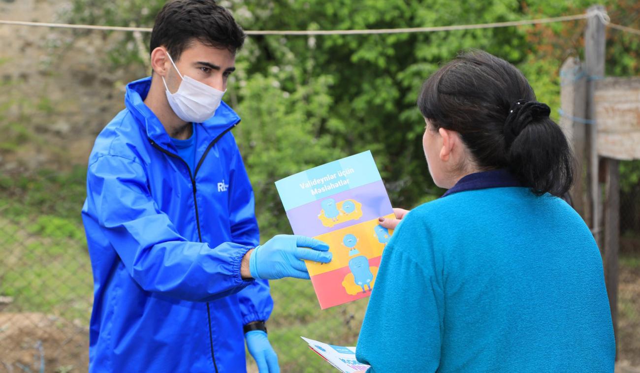 Une femme tend une brochure à un jeune homme portant des gants et un masque de protection.