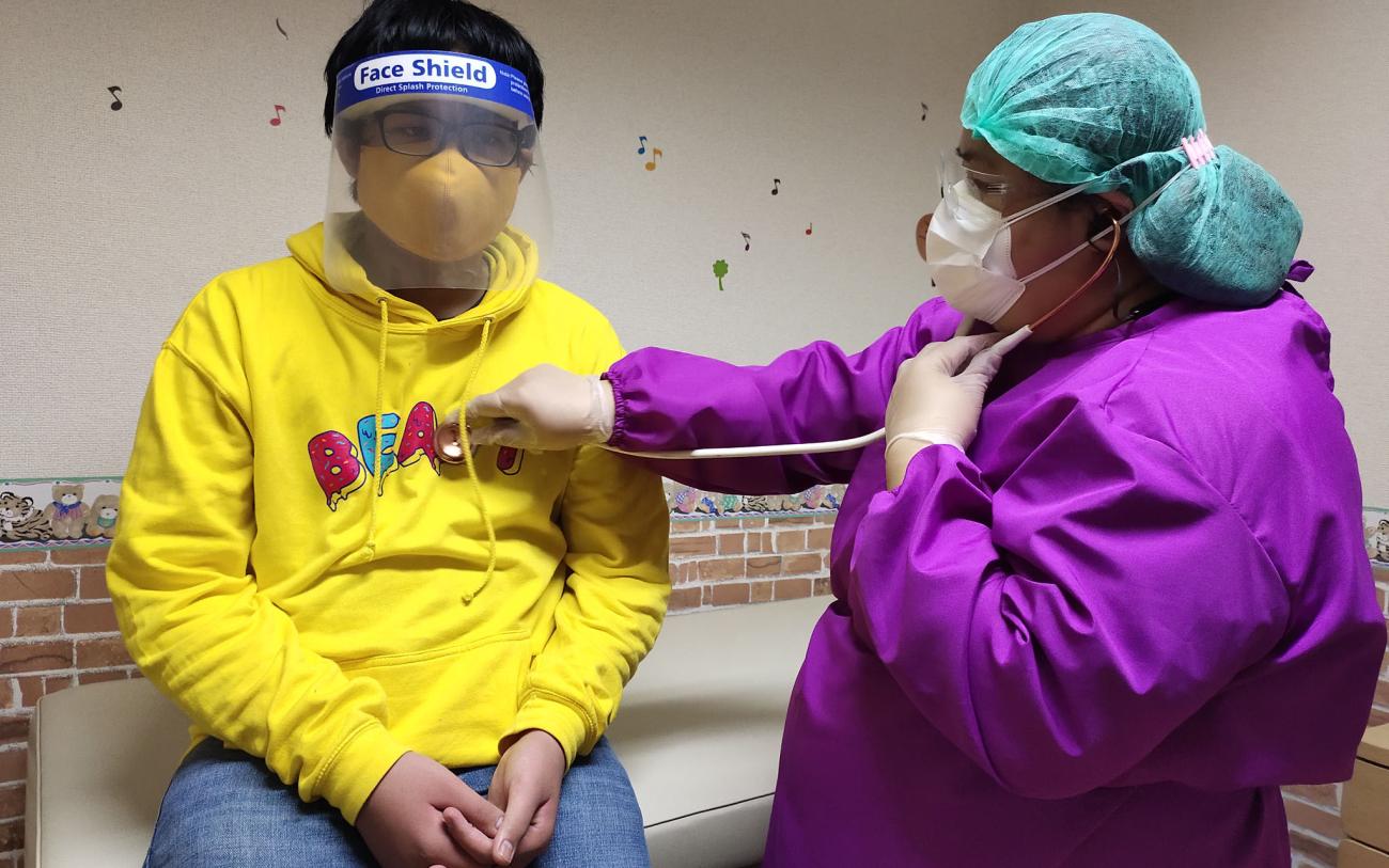 Une femme-médecin ausculte un jeune garçon dans un centre de soins. Le garçon porte un masque et une visière de protection, tandis que la médecin est vêtue d’une tenue de protection et porte un masque et une charlotte.