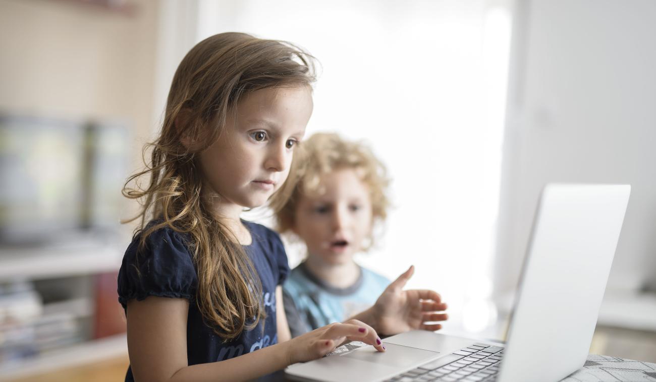 Une petite fille utilise un ordinateur portable. Son petit frère se tient près d’elle et observe ce qu’elle fait avec intérêt.