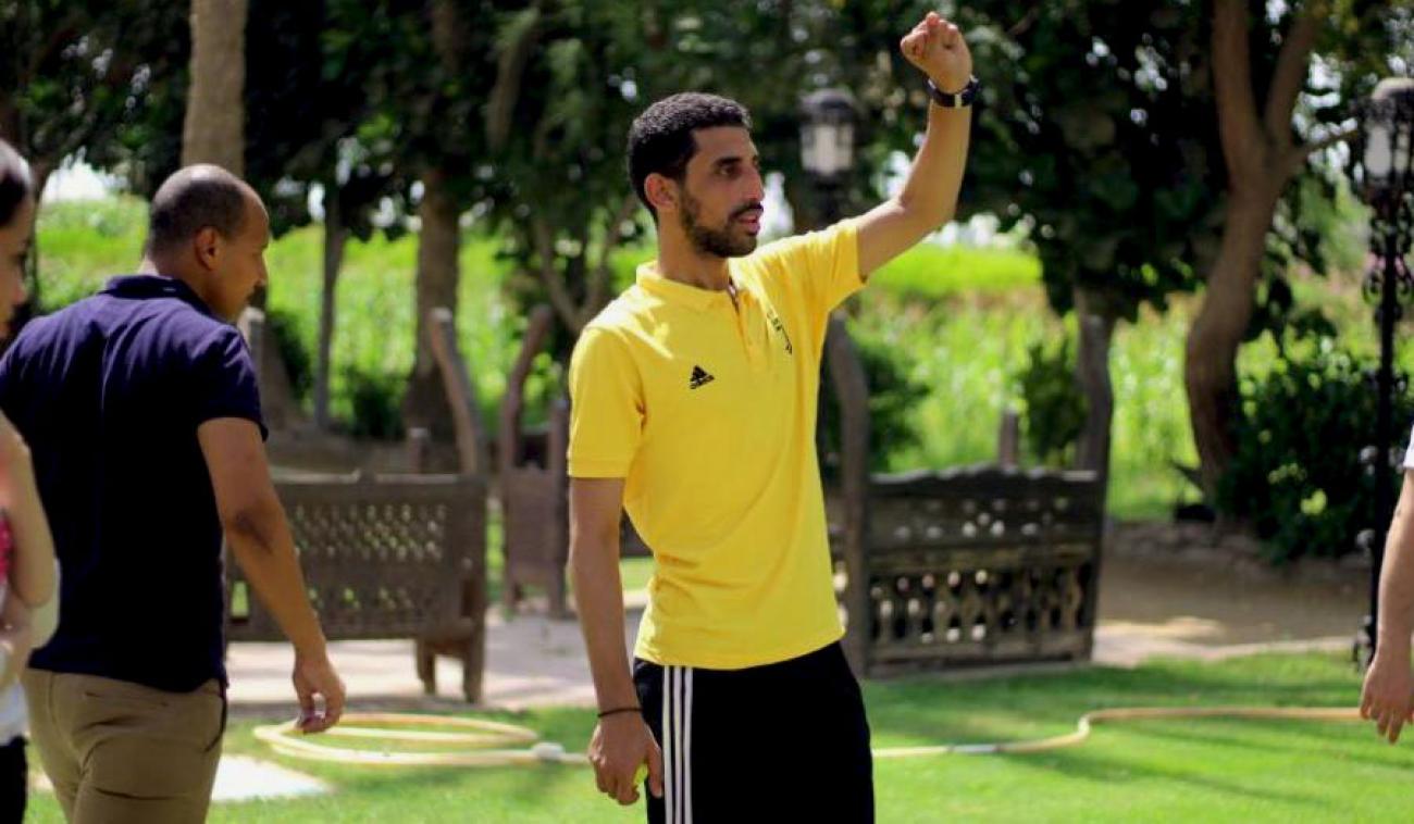 Mohamed ElKholy está parado en el centro de la imagen, en el fondo se aprecia un parque con grama. Mohamed está usando un pantalón de chándal negro y una camiseta amarilla. Levanta el brazo izquierdo para llamar la atención a otra persona.