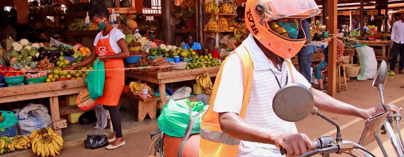 Un conductor de SafeBoda sale del mercado en un ciclomotor para entregar comida. Lleva una máscara y un casco protector. Al fondo, una mujer llena una bolsa de frutas en uno de los muchos puestos de frutas y verduras del mercado.