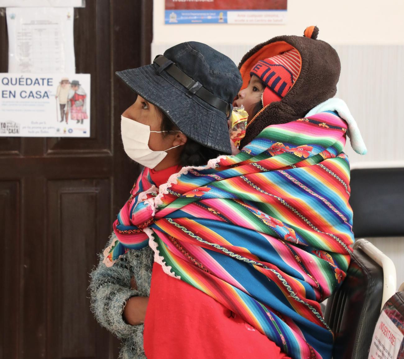 Foto de una mujer boliviana, con un fular tradicional con el cual carga en su espalda a un niño pequeño, y ambos están esperando en un pasillo de un centro de salud local.