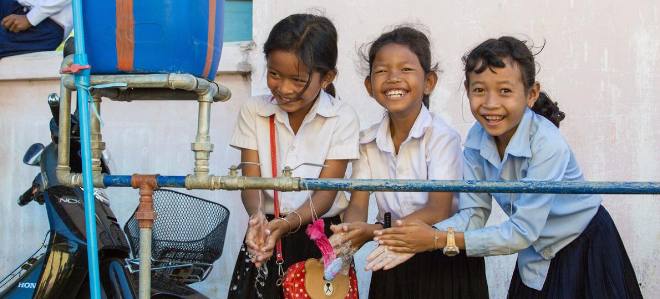 Las niñas de una escuela en Camboya, vestidas con sus uniformes escolares, se lavan las manos con agua de las instalaciones de "Agua, Saneamiento e Higiene para Todos" de una escuela. Las tres se ríen mientras se lavan las manos y dos de ellas miran a la cámara.
