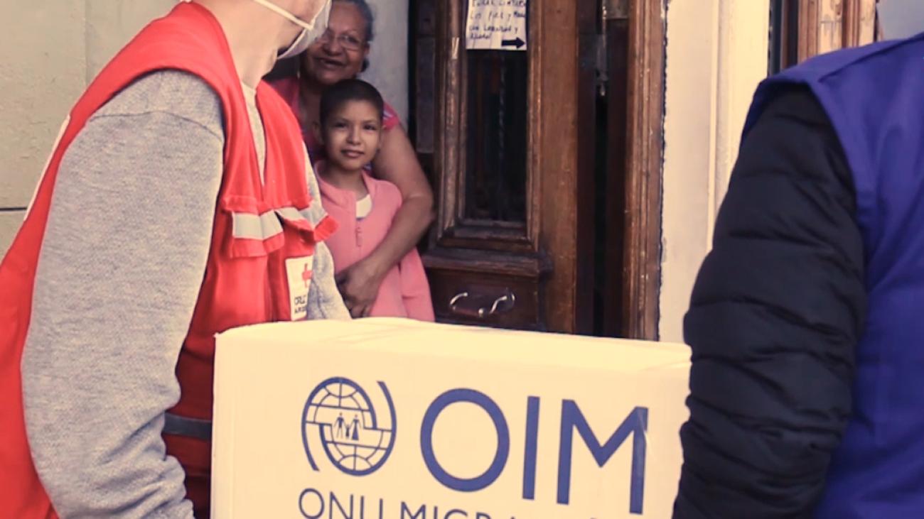 Deux membres du personnel de l'OIM viennent remettre un carton de fournitures à une famille. Une femme âgée et un jeune garçon se tiennent sur le seuil de la porte pour les accueillir.
