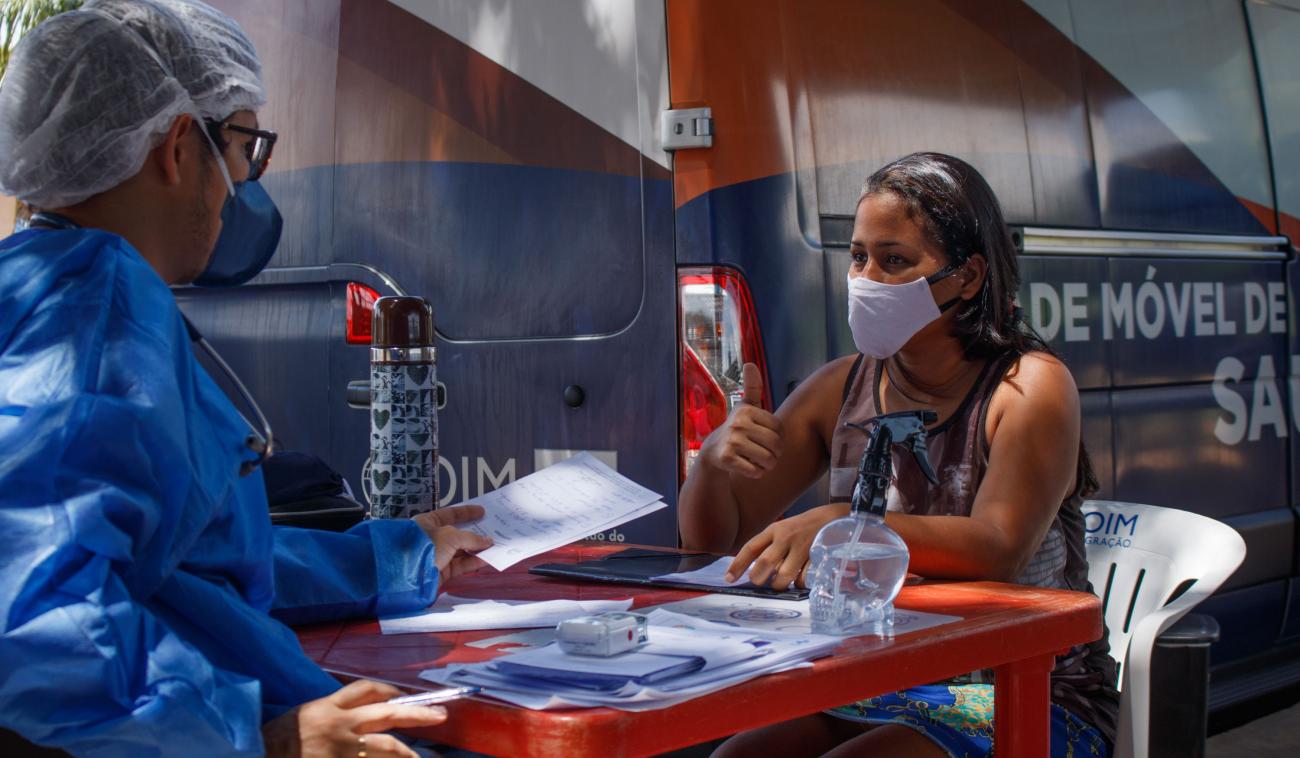 Un membre du personnel de l’OIM portant un masque et un équipement de protection individuelle est assis à une table avec une femme portant elle aussi un masque de protection. L’homme remet un document à la femme, qui lève le pouce.