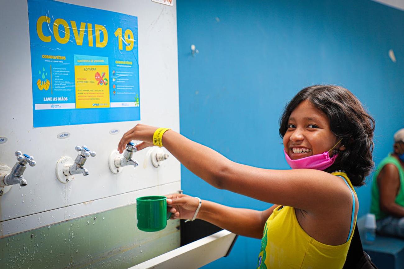 A young Venezuelan girl wearing a mask under her mouth happily smiles as she prepares to fill her mug at a drinking water station at a shelter. 