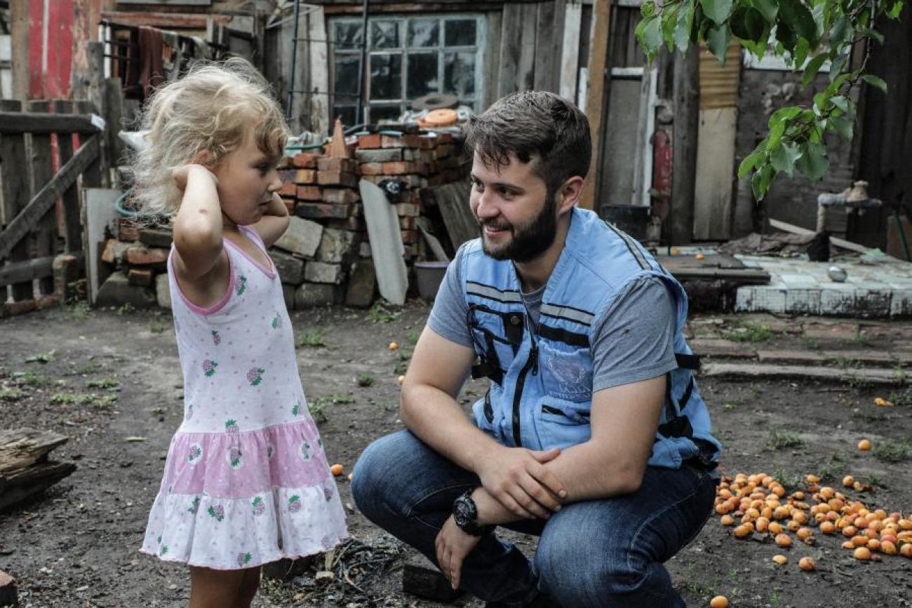 Un miembro del personal de la ONU está agachado y sonriendo mientras habla con una niña pequeña, que levanta los brazos al hablar. El hombre y la niña aparecen en el patio de una casa, en Ucrania.