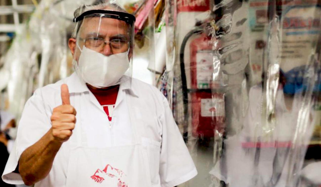 Un comerciante que lleva una visera protectora y una bata blanca de laboratorio se encuentra cerca de un puesto de comida protegido por láminas de plástico. Mira a la cámara con un pulgar apuntado hacia arriba, de forma afirmativa.