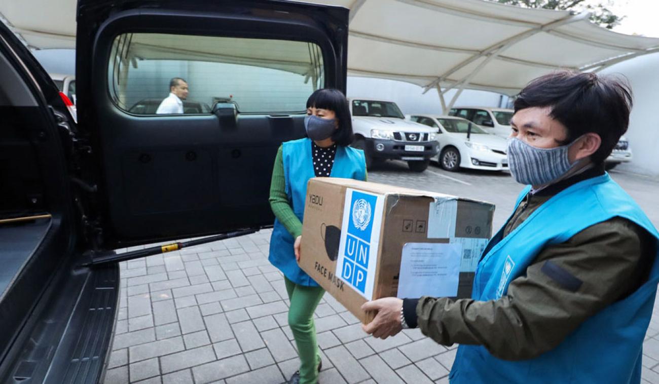 Dans un parking, un homme et une femme membres du personnel de l'ONU s'apprêtent à déposer un grand carton de fournitures offertes par le PNUD dans le coffre d'un véhicule. Chacun d'eux porte un masque de protection respiratoire.