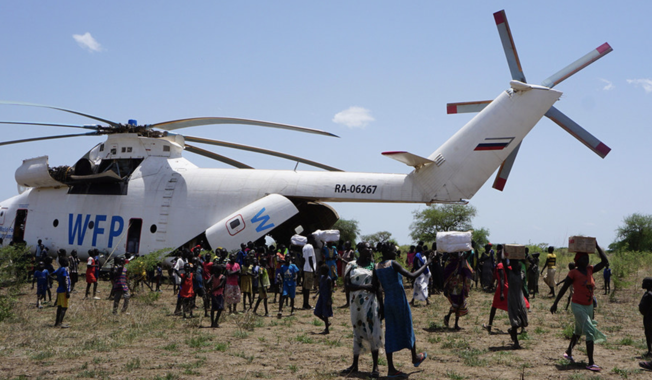 Un helicóptero del Programa Mundial de Alimentos entrega suministros esenciales a la población de Udier, Sudán del Sur. Muchas personas, incluidos niños, están reunidas alrededor del helicóptero. Entre ellos, se ve a mujeres alejándose del vehículo aéreo de transporte, con cajas y otros tipos de paquetes en la cabeza.