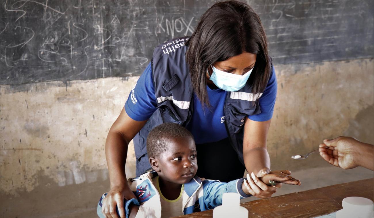 Au Zimbabwe, dans la salle de classe d’une école locale, un membre féminin du personnel de l'OMS portant un masque de protection aide un jeune garçon à tendre la main pour qu'on lui donne un médicament. Une personne hors-champ lui tend le comprimé dans une cuillère. En arrière-plan, on voit un tableau d’école.