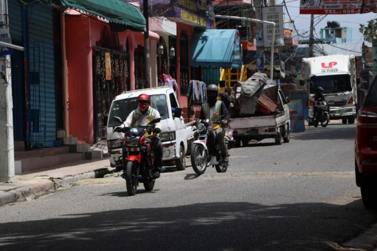Scène de la vie citadine où l’on voit des motocyclistes portant chacun un masque de protection et un casque de sécurité circuler dans une rue et plusieurs fourgonnettes stationnées sur le côté.