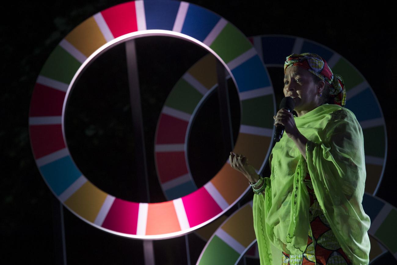 La Vicesecretaria General de las Naciones Unidas y la Presidenta del Grupo de las Naciones Unidas para el Desarrollo Sostenible habla, en el escenario, por un micrófono mientras está de pie, junto a grandes recortes de ruedas en el fondo, que representan el logo de los ODS.
