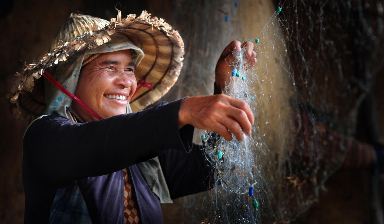 Una mujer, sonriente, seca una red de pesca. La foto se titula "Happy work" y su protagonista luce indumentaria que le protege los brazos (y el cuello). Lleva puesto un sombrero de paja, de ala ancha.